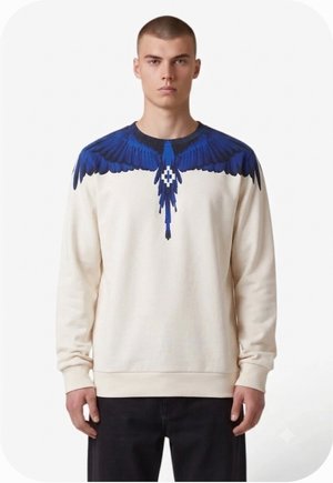 Giovane uomo che indossa una felpa color crema con un motivo di ali e coda di uccello blu che si estende sulle spalle e sul petto.