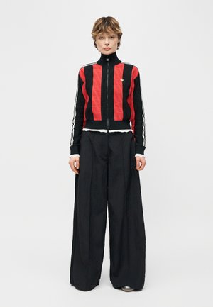 Tânăr cu păr scurt, purtând o jachetă cu fermoar roșu și negru în dungi și pantaloni negri largi, stând în picioare pe un fundal simplu.