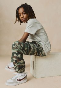 Grau kurzärmeliges Hemd, Camouflage-Hose mit Taschen und weiße Sneaker mit roten Akzenten, sitzend auf einem hellen Block vor einem neutralen Hintergrund.