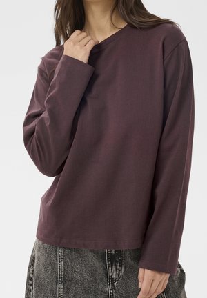 Femme portant une chemise à manches longues violet foncé et un jean noir délavé, une main touchant légèrement l'encolure.