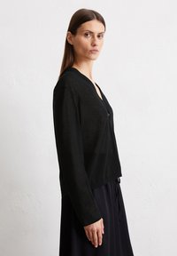 Schwarze, durchsichtige Strickjacke mit tiefem V-Ausschnitt, Knopfdetail, langen Ärmeln und offener Front. Strukturierter Stoff umschmeichelt sanft ein dunkles Kleid.