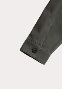 Green cotton shirt cuff with a black button and stitching details, featuring a straight edge and textured fabric.