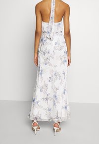 Bakifrån av en kvinna som bär en vit halterneck-blommig maxiklänning med blå och lila blommor samt vita högklackade sandaler.