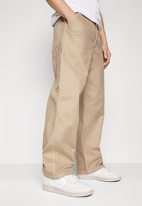 Beiga cargobyxor med en avslappnad passform, tillverkade av slitstarkt tyg, med sidofickor och kontrasterande sömdetaljer. Bärs med vita sneakers.