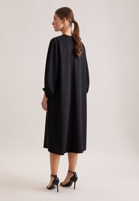 Schwarzes, locker sitzendes Kleid mit langen, voluminösen Ärmeln und rundem Ausschnitt. Glatte Textur und Midi-Länge, kombiniert mit schwarzen High Heels.