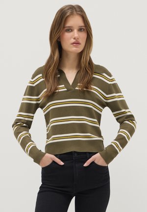 Jeune femme aux longs cheveux portant un pull à col en V rayé vert olive, blanc et jaune, et un pantalon noir, les mains dans les poches, fond neutre.