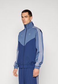 Jachetă sport albastră cu guler înalt, design colorat în chevron în nuanțe de bleumarin și albastru deschis, având trei dungi albe pe mâneci.
