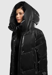 Zwarte pufferjas met een hoge, oversized capuchon, glanzende afwerking, ritssluiting aan de voorkant en zijzakken, met een doorgestikte textuur en merkdetails.