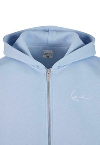 Lys blå zip-up hettegenser med en myk tekstur, sølvglidser og en hvit brodert logo på venstre side. Har en romslig hette.
