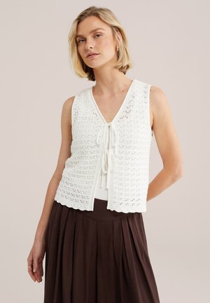 Heledate juustega naine, kes kannab esiküljelt seotud valget varrukateta heegeldatud vestit valge topi ja pruuni volditud seelikuga.