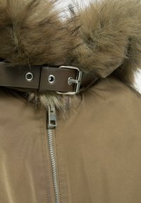 Tanja izolirana jakna s kapuco iz umetnega krzna, rjav umetni usnjeni pas s srebrno sponko in kovinskim detajlom zadrge. Gladka zunanja tekstura.