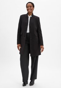 Cappotto nero con colletto alto, caratterizzato da una chiusura con bottoni e tasche laterali. Indossato sopra una camicia bianca e pantaloni grigi a righe.