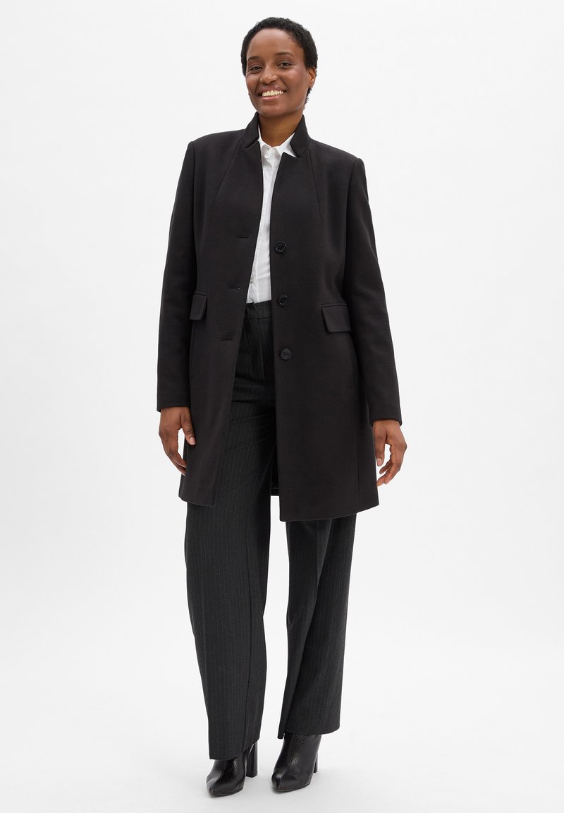 Cappotto nero con colletto alto, caratterizzato da una chiusura con bottoni e tasche laterali. Indossato sopra una camicia bianca e pantaloni grigi a righe.
