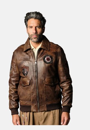 Homme aux cheveux poivre et sel portant une veste en cuir marron avec des patches et une fermeture éclair, debout devant un fond uni.