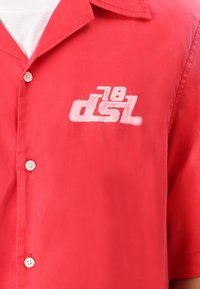 Camisa roja de manga corta en tejido ligero. Presenta un logo blanco impreso, cuatro botones blancos y un collar puntiagudo.