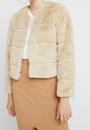 Nainen, jolla on päällään beige keinoturkistakki valkoisen topin ja vaaleanruskean ribbineulotun hameen päällä, kuvattu kaulasta lanteisiin.