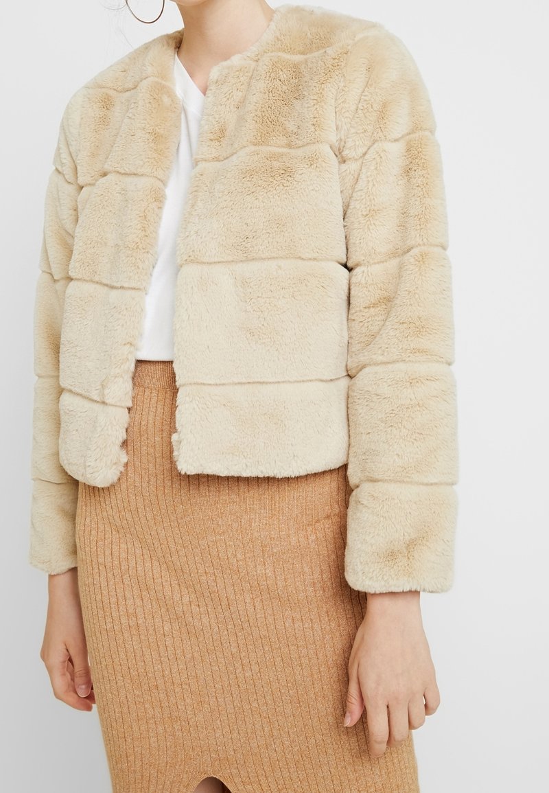 Femme portant une veste en fausse fourrure beige sur un haut blanc et une jupe côtelée marron clair, visible du cou aux hanches.
