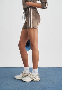 Γυναίκα που φοράει καφέ βελουτέ σορτς με λευκές ρίγες, λευκές κάλτσες και μπεζ αθλητικά παπούτσια Nike, στέκεται σε μπλε χαλί κρατώντας μπλε καπέλο.