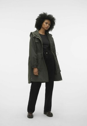 Olivegrüner langer Parka mit Kapuze, Kordelzug in der Taille und Vordertaschen, kombiniert mit einem schwarzen Oberteil und lockeren schwarzen Hosen. Braune klobige Schuhe.