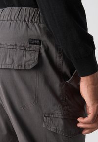 Pantalon cargo gris avec une ceinture élastique, possédant des poches latérales et une étiquette. Le tissu semble lisse et léger avec des coutures visibles.