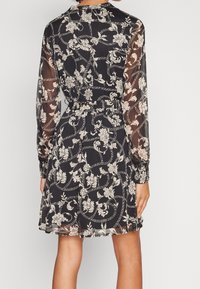 Kvinna klädd i en svart blommig klänning med vita vinmönster, genomskinliga långa ärmar och åtdragen midja, sedd bakifrån.