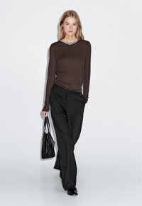 Brauner langärmliger Strickoberteil, über ein graues Shirt getragen, kombiniert mit lockeren schwarzen Hosen und schwarzen Schuhen. Das Model trägt eine schwarze Tasche.