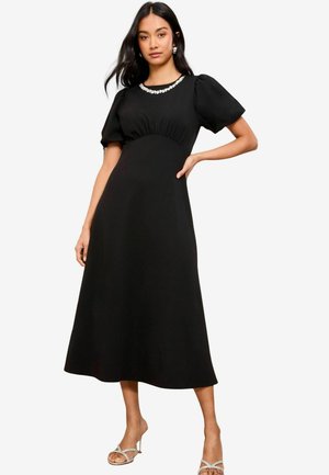 Abito midi nero con maniche a sbuffo, corpetto arricciato e scollatura impreziosita. Tessuto morbido con una silhouette fluida, abbinato a sandali con tacco.