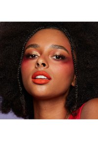 Närbild av en modell med mörkt, lockigt hår, djärv orange läppstift och ögonskugga som blandar guld- och röda nyanser på en mjuk bakgrund.