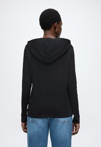 Μαύρο μακρυμάνικο φούτερ με κουκούλα και κεντρική ραφή στην πλάτη, μαλακή υφή και χαλαρή γραμμή, συνδυασμένο με ανοιχτόχρωμο τζιν παντελόνι.