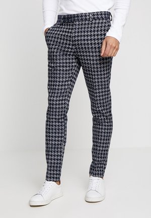 Mężczyzna ubrany w dopasowane spodnie w czarno-szary wzór pepitki, białe trampki i biały, długi rękaw, ręce w kieszeniach.