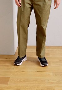 Pantaloni dritti color khaki scuro abbinati a sneakers bianche e nere, con una texture liscia e un design minimalista.