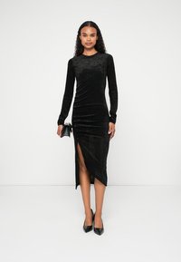 Femeie îmbrăcată într-o rochie midi neagră din catifea cu mânecă lungă și o fanta pe coapsă, pantofi cu toc negri și ținând o poșetă mică neagră.