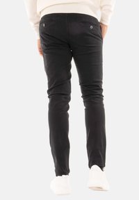 Pantaloni neri a sigaretta in cotone con una texture liscia. Presentano due tasche posteriori e polsini elastici alle caviglie, abbinati a scarpe bianche.