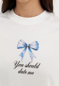 Biała koszula z niebieską grafiką w kształcie kokardy i czarnym kursywą napisem "Powinieneś się ze mną umówić" na klatce piersiowej, pokazana od szyi do górnej części torsu.