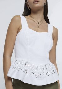 Bílý peplum top s čtvercovým výstřihem, texturovanou látkou a vyšívanými detaily na okraji. Ozdobený vrstvami zlatých náhrdelníků.