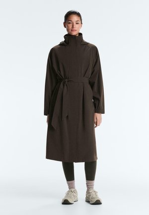 Femme portant un manteau brun foncé ceinturé jusqu'aux genoux, des leggings noirs, des chaussettes grises et des baskets beiges, debout devant un fond clair uni.