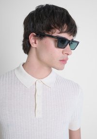 Polo a maniche corte in tessuto bianco sporco, con colletto classico e placket a tre bottoni. Il modello indossa occhiali da sole rettangolari scuri.