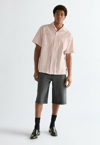 Κόκκινο και λευκό ριγέ πουκάμισο με κοντά μανίκια, δύο τσέπες στο στήθος, σε συνδυασμό με σκούρες τζιν βερμούδες μέχρι το γόνατο και μαύρα μοκασίνια, συνοδευόμενα από μαύρες κάλτσες.