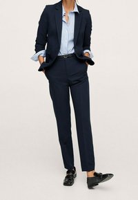 Kobieta w granatowej marynarce i spodniach, z niebieską koszulą w paski wpuszczoną do środka, z rękami w kieszeniach, stojąca z lekko uniesioną jedną stopą.