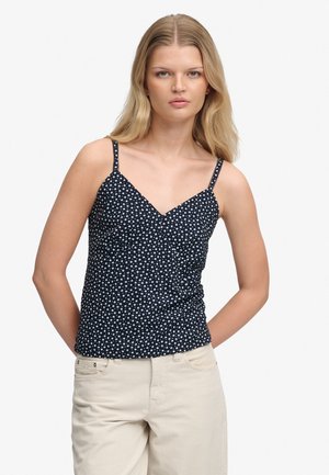 Femme blonde portant un haut noir sans manches avec un imprimé floral blanc et un pantalon beige, debout devant un fond blanc.