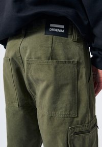Tummanvihreät cargo-housut, joissa on useita taskuja, vetoketjullinen tasku ja musta DRDENIM-tunnus vyötäröllä. Sujuva ja kestävä kangas.
