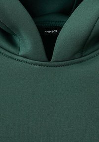 Tela verde y suave con una textura lisa y un diseño con capucha. Presenta un escote en forma de V y sutiles detalles de costura. Etiqueta de la marca visible.