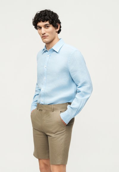 Άνδρας που φοράει ανοιχτό μπλε πουκάμισο με κουμπιά σε γαλάζιο χρώμα, μπαίνει μέσα σε μπεζ ραμμένα σορτς, στέκεται με τα χέρια στις τσέπες σε απλό φόντο.