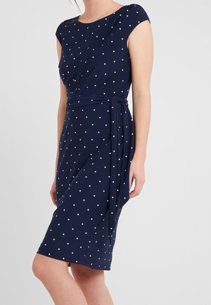 Vrouw draagt een marineblauwe jurk tot op de knie met witte polkadots, kapmouwen en een taillestrop, staand tegen een effen achtergrond.