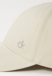 Cappello beige in tessuto testurizzato, con logo "ck" in argento sul davanti. Visiera curva e dettagli cuciti lungo le cuciture.