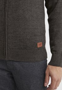 Närbild av en person som bär en mörkgrå stickad tröja med dragkedja och en liten brun etikett nära midjan samt mörkgrå byxor.