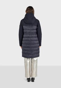 Abrigo acolchado azul marino con capucha, con secciones de acolchado horizontal y parte superior lisa. Combinado con pantalones metálicos de pierna ancha.