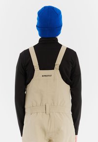 Persoon in beige tuinbroek met het logo "PROTEST" op de achterkant en een zwart longsleeve shirt, met daarop een felblauwe gebreide muts, met de rug naar ons toe.
