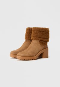 O pereche de botine din suede bej cu tivuri din blană tăiată, tocuri pătrate și talpa marcata cu "LOVE MOSCHINO."