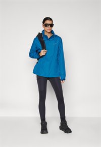 Blå vanntett jakke med hette, kombinert med svarte leggings og tursko. Har en svart skulderryggsekk og mørke solbriller.